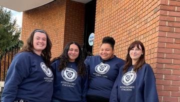 Four female students wearing Navy blue sweatshirts pose together in front of a brick wall