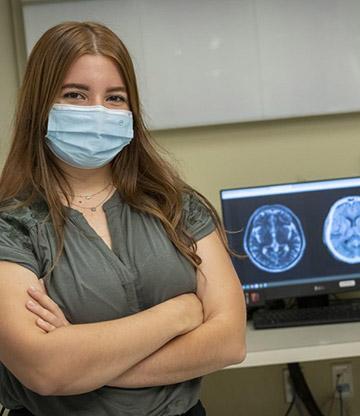 Isabella wears a green shirt and a mask, while standing in front of an MRI scan