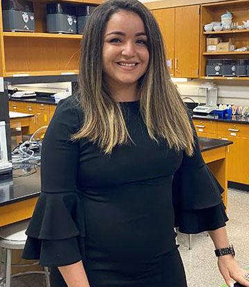 Paola wears a black dress and stands in a science lab