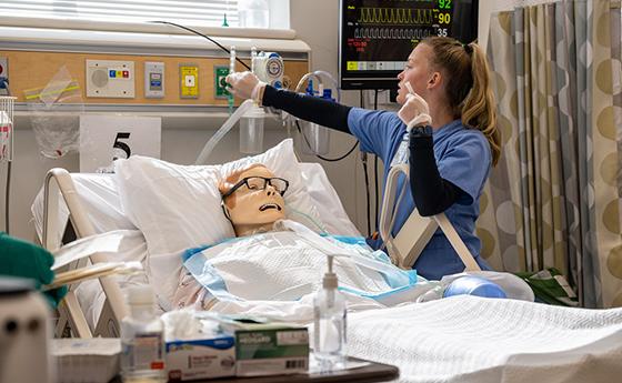 Female student in light blue scrubs uses hospital equipment with a manikin