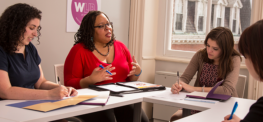 Four women in a professional setting engaged in discussion and taking notes